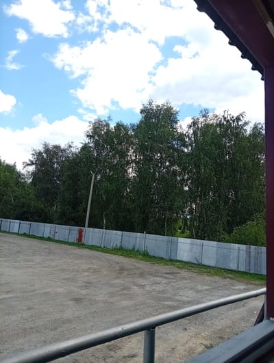 This is an outdoor, eye-level shot of a gravelly area, possibly a training ground or yard, bordered by a corrugated metal fence. In the background, a line of dense deciduous trees, likely birch, forms a natural backdrop. Above the trees, a bright blue sky is dotted with white, fluffy clouds. The scene is framed on the right by the edge of a dark red structure, possibly a building or overhang. A metal railing runs horizontally across the foreground, obscuring part of the ground. The overall impression is one of a rural or semi-rural location, possibly on the outskirts of the city of Kushva, Russia.