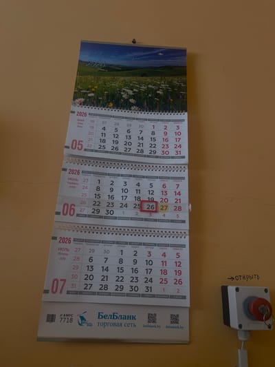 The image shows a wall calendar for the year 2026, featuring landscapes of Belarus on each monthly page. The calendar is hanging on a yellow wall, with a red emergency stop button and a label with the Russian word "открыть" (open) visible in the upper right corner.
The calendar is designed in a panoramic format, with three months displayed side-by-side. The visible months are May, June, and July. Each month has its own corresponding landscape image. The May page features a field of daisies and grass under a blue sky. The June and July pages are partially obscured but appear to continue the theme of Belarusian nature.
The calendar grids are printed in Cyrillic and Latin alphabets, indicating a bilingual audience. The days of the week are listed as "НЕДЕЛЯ" (Sunday), "ПОНЕДЕЛЬНИК" (Monday), "ВТОРНИК" (Tuesday), "СРЕДА" (Wednesday), "ЧЕТВЕРГ" (Thursday), "ПЯТНИЦА" (Friday), and "СУББОТА" (Saturday). The months are labeled in Russian ("МАЙ", "Июнь", "Июль") and English ("May", "June", "July").
Promotional text and logos for "БелБланк" (BelBlank) and "торговая сеть" (trading network) are visible on the bottom right of the calendar, along with the website addresses "belblank.by" and "datamark.by." A barcode and the number "7718" are also present.
The overall scene suggests an office or public space in Masty, Belarus, where the calendar serves both as a timekeeping tool and a decorative element showcasing the country's natural beauty. The lighting is bright, indicating it's daytime.
