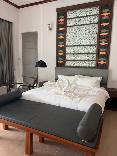 The image depicts a hotel bedroom in Ban Kao Dua, Thailand. The room is furnished with a king-size bed with white linens, two white towel swans placed on the pillows, and a gray upholstered headboard. A dark gray bench with a wooden frame sits at the foot of the bed. The headboard is adorned with a decorative panel featuring floral fabric and geometric patterns in green, orange, and brown. To the left of the bed, a gray armchair and a small table are visible, along with gray curtains covering a window. A black pendant light hangs from the ceiling. On the right side of the bed, a dark wooden nightstand is present. The walls are painted white, and the floor is tiled with light-colored wood-patterned tiles.