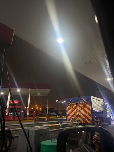The image shows a gas station at night in Lurín, Peru. The station has a red and white canopy illuminated by bright lights. Fuel pumps are visible, displaying Petroperú branding. A truck with a blue and white container and a red and yellow chevron pattern is parked nearby. The rear of the truck has directional arrow lights. The license plate BMA-433 is visible on the truck. The view is from inside a car, with a side mirror visible showing a hand holding a cell phone.