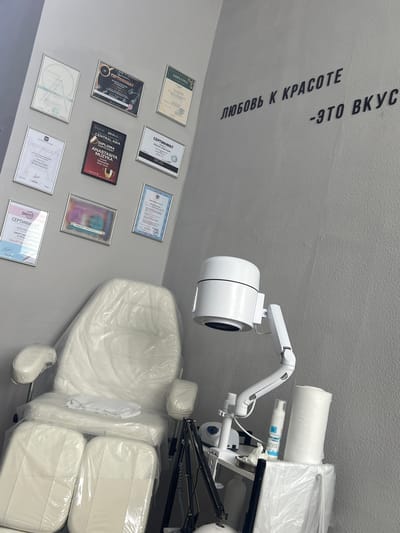 The image shows a clean, modern interior of a beauty salon. On the wall, there are several framed certificates and diplomas, suggesting that the salon is professionally certified and its staff are skilled. The diplomas are in various languages, indicating an international clientele or training. The text on the wall, "ЛЮБОВЬ К КРАСОТЕ - ЭТО ВКУС" (Love for beauty is taste), conveys the salon's philosophy.
In the foreground, a white beauty chair, covered in plastic for hygiene, is prominently displayed. Beside it, a white trolley holds essential beauty equipment, including a lamp with adjustable arm and a roll of paper towels. A bottle of product with Cyrillic text is also visible on the trolley. The overall impression is one of a well-equipped and meticulously maintained beauty salon, likely in a commercial or residential area of Stepnogorsk city administration, Kazakhstan, catering to clients seeking aesthetic services.