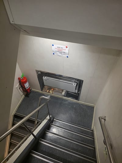 The image shows a top-down view of a dimly lit staircase with dark grey flooring and metal railings. On the wall to the left, there is a red fire extinguisher. Above the stairs, a sign is attached to the wall with two red pushpins and one blue pushpin. The sign reads: "KEEP ALL "ACCOMODATION DOORS" CLOSED ALL THE TIME ENG. DEPT." In the wall above the stairs, there is a recessed area containing a white and silver HVAC unit. The walls are light-colored and appear to be paneled. The overall scene suggests an interior, possibly on a ship or in a utilitarian building, given the presence of a fire extinguisher and a technical sign. There is no indication of the city of Brindisi, Italy, in the image.