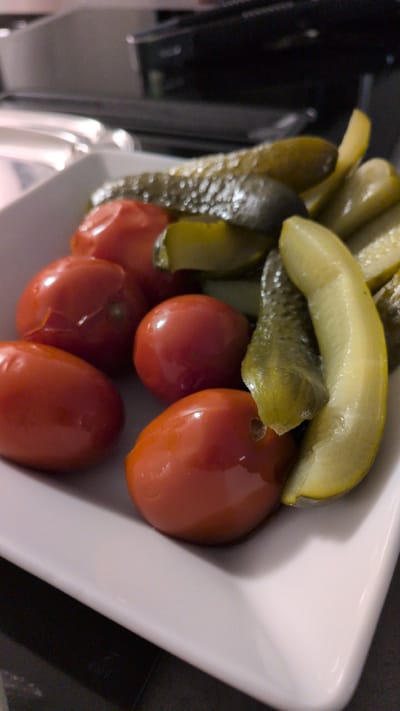 This is a close-up, low-angle, slightly tilted shot of a white rectangular dish filled with pickles and cherry tomatoes. The pickles appear to be dill pickles, cut into spears, with some whole smaller pickles visible. They are a mottled green and yellow color. The cherry tomatoes are small, round, and bright red, with a glossy sheen. They are nestled amongst the pickles. The dish is partially filled, and the contents are arranged in a way that suggests they are a garnish or appetizer.

The setting appears to be indoors, possibly a restaurant or home kitchen, given the clean white dish and the dark background which might be a countertop or table. There is no indication of the time of day, weather, or any specific emotions conveyed. No text is visible in the image. The overall impression is a simple, fresh presentation of pickled vegetables and tomatoes. The lighting is somewhat dim, creating soft shadows within the dish. The resolution of the image is moderate, allowing for a good view of the textures of the food.