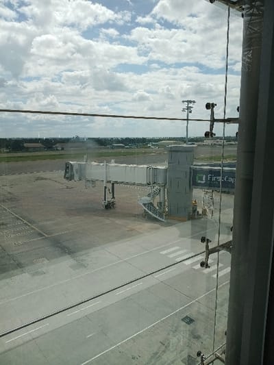 The image displays an elevated view from inside an airport terminal at Robert Gabriel Mugabe International Airport in Harare, Zimbabwe. The sky above is partly cloudy with numerous white cumulus clouds against a blue background.

In the immediate foreground, a large glass window pane shows reflections, thin horizontal wires, and vertical metal support structures. The midground features a grey concrete tarmac marked with white painted lines and striped patterns. A light-colored passenger boarding bridge (jet bridge) is positioned centrally, extended but not connected to an aircraft. It includes a mobile base, a retractable walkway section, and an attached circular metal staircase descending to the tarmac. Adjacent to the jet bridge's fixed support column, a blue structure with "First Capital" text in white lettering is visible. A small, dark ground support vehicle is situated directly beneath the jet bridge's mobile section. Two indistinct human figures are present near the base of the jet bridge or the 'First Capital' structure.

The background reveals a flat expanse of green ground or grass, leading to a distant horizon line where low-rise buildings and scattered green vegetation are discernible. Tall metal poles equipped with multiple light fixtures are visible in the middle distance on the airport grounds. No specific actions or interactions are clearly observed; the scene appears static.