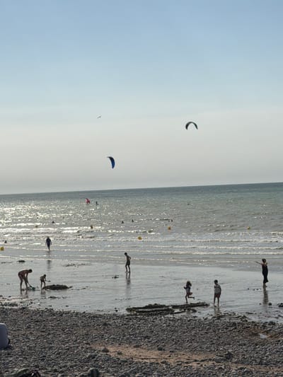 The image depicts a bright, sunny day at a beach in Hautot-sur-Mer, France. The scene is divided into two distinct areas: a vast expanse of pale blue sky and a shoreline with the ocean. The sky is clear, with a few kites visible. One kite, dark in color, is seen in the upper left quadrant. Another, a vibrant blue, is further down and to the right. A third, smaller kite or object, appears in the far distance.

The shoreline is a mix of wet sand and pebbles, reflecting the sunlight intensely. Several people are scattered along the beach and in the shallow water. In the upper right section of the image, two figures are walking on the wet sand, their shadows elongated. Below them, another person is walking, their silhouette clearly defined. Further down, a person appears to be kneeling. Towards the bottom of the image, a person is seen standing, possibly near the water's edge, with another individual in the distance. The wet sand and shallow water create a shimmering effect due to the sunlight. Small, yellow buoys are dotted along the edge of the water. The overall atmosphere suggests a leisurely day at the beach, with activities like kite flying and simply enjoying the seaside. The time of day appears to be late afternoon, given the long shadows.