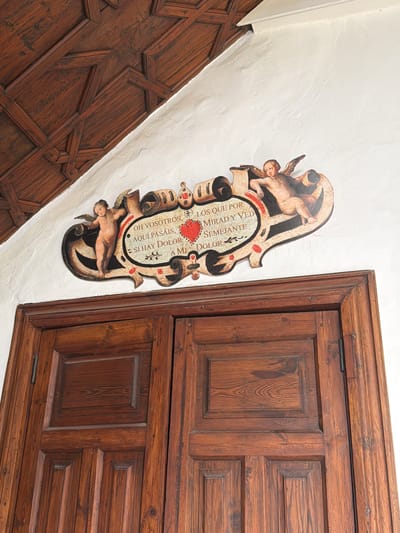 The image shows a close-up view of a wooden door and an ornate plaque on a white wall. The door is made of dark, aged wood with visible grain and knot patterns. It appears to be a double door, with the right door slightly more visible. The door features raised panels, a common decorative element in traditional woodworking.
Above the door, mounted on the white wall, is a decorative plaque. The plaque is elaborately designed with scrollwork and two cherubic figures, one on the left and one on the right. These figures are depicted in a classical style, with wings and child-like bodies. The plaque itself has a cream-colored background with intricate black and red detailing. In the center of the plaque, there is a red heart symbol, and around it, there are inscriptions in what appears to be Spanish. One section of the inscription reads "OH VOSOTROS, LOS QUE POR AQUI PASÁIS, MIRAD Y VED SI HAY DOLOR SEMEJANTE A MI DOLOR." This translates to "OH YOU WHO PASS HERE, LOOK AND SEE IF THERE IS PAIN LIKE MINE."
The wall behind the plaque is textured and painted white, contrasting with the dark wood of the door and the rich colors of the plaque. Above the wall, a section of a dark wooden ceiling is visible. This ceiling has a geometric, coffered design, suggesting an older architectural style. The lighting in the image suggests an indoor environment, possibly a historical building or a well-preserved interior space. The overall scene conveys a sense of history and artistic craftsmanship. The location of La Cuesta, Spain, is not directly evident from the visual details of the image itself, but is provided as contextual information.