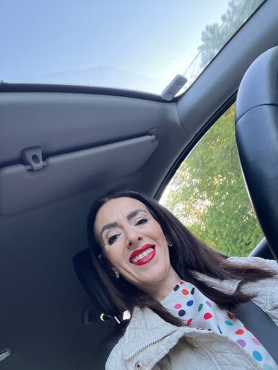 A woman with long dark hair, dark eyebrows, and bright red lipstick is captured in a close-up photo inside a car. She is smiling broadly, showing her teeth, and appears cheerful. She wears an off-white quilted jacket over a white top adorned with colorful polka dots (red, orange, green, blue, black, pink). A seatbelt crosses her chest.
The scene is set inside a vehicle, with elements of the car's interior visible. A black steering wheel is in the upper right, and a gray sun visor is on the left side of the frame. Through the car's windows, bright green trees and a clear sky are visible, indicating it is daytime.
A small, white rectangular tag is affixed to the windshield, near the left A-pillar. This tag features a barcode and some text. Clearly visible are the numbers "10280456," the abbreviation "LUXBG" (for Luxembourg), and the French phrase "CONTROLE TECHNIQUE" (Technical Inspection), suggesting it is a vehicle inspection sticker from Luxembourg. The overall setting implies the woman is driving or parked in Grevenmacher, Luxembourg.