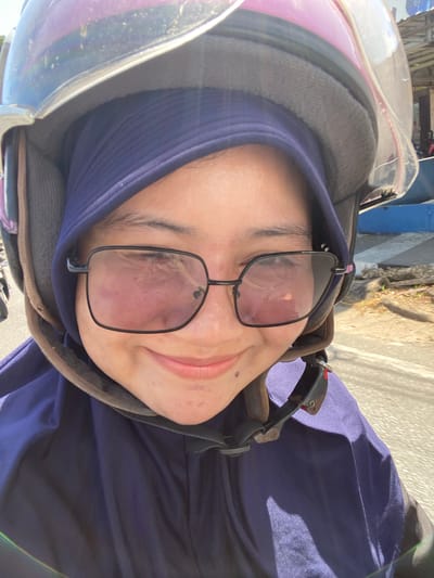 A woman wearing a purple hijab and a motorcycle helmet takes a selfie. She is wearing glasses with black frames. The background shows a blurred outdoor scene with a road and buildings. The scene is likely in Keraton, Indonesia, given the contextual information.