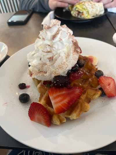 A white plate displays waffles topped with whipped cream, strawberries, blueberries, and blackberries. The waffles are golden brown and appear crisp. The whipped cream is piled high and dusted with cocoa powder. Sliced strawberries are arranged around the waffle, adding a vibrant red color. Blueberries and blackberries are scattered among the other ingredients. A wrap is visible in the background on a smaller black plate.