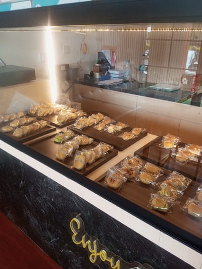 A display case filled with various wrapped pastries, arranged on dark wooden trays. The pastries appear to be different types of bread and buns. The case has a marble-patterned lower section. At the bottom, a yellow neon sign spells "Enjoy". Behind the pastries, the display case reveals a countertop with water bottles and other items. A bright glare obscures the left side of the case.