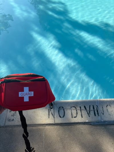 A red lifeguard fanny pack with a white cross and the word "GUARD" on it rests next to a swimming pool in Edison, New Jersey, United States. The pool's edge has the words "NO DIVING" painted on it. Sunlight reflects off the water in the pool. A black twisted cord is attached to the fanny pack.