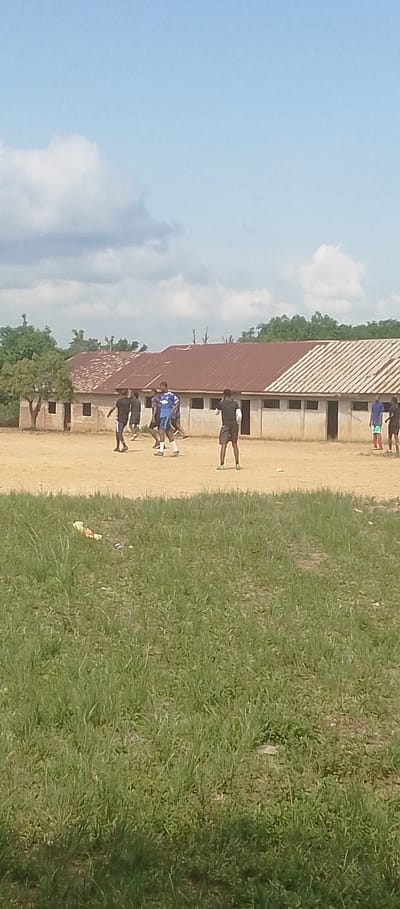 A group of young men are engaged in a soccer game on a dusty field in front of a long, single-story building with a rust-colored, corrugated metal roof. The building has multiple windows and doors, and appears to be made of concrete blocks. Trees and vegetation surround the building, with a patch of tall green grass in the foreground of the image. The sky is a bright, clear blue with a few scattered white clouds. The sun appears to be high in the sky, suggesting it is midday. The players are dressed in athletic wear, with some wearing shorts and t-shirts, and others in soccer jerseys. The scene appears to be set in Achara Ndiagu, Nigeria, given the architectural style of the building and the surrounding natural environment. The overall atmosphere is casual and recreational, with the men focused on their game. There is no visible text in the image.