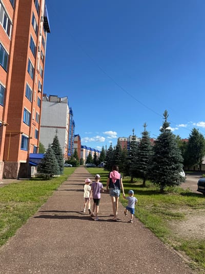 The image shows three young people walking on a paved path next to a grassy area. The person in front is wearing a pink shirt and dark shorts, with a pink backpack. To their right, another person in a purple shirt and dark shorts walks alongside. A third person, wearing a striped shirt, blue shorts, and a blue cap, walks slightly ahead of them. All three appear to be children or young teenagers.
Behind the grassy area, several tall evergreen trees are visible, casting long shadows on the path. Beyond the trees are residential buildings, some made of red brick with many windows, and others with lighter facades and blue-tinted windows. The sky is a clear, bright blue with a few white clouds. The sun is high in the sky, indicating it is likely midday. The overall scene suggests a warm, sunny day in a residential area, possibly a park or courtyard. There are no visible emotions conveyed, but the activity appears to be casual walking or strolling.
The image is oriented vertically, with the sky occupying the upper portion and the ground and buildings in the lower portion. The paved path curves gently to the right as it recedes into the distance. There is a car parked on the side of a road visible in the background, to the left of the evergreen trees. The location context is identified as Mariinsky, Russia. No visible text is present in the image, except for what might be on the license plate of the car in the background, which is too blurry to read.