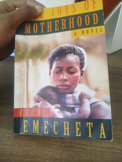 A dark-skinned hand is positioned at the top-left, holding a copy of the paperback novel "THE JOYS OF MOTHERHOOD" by Buchi Emecheta. The book's cover features a close-up image of a Black woman with braided hair and a light head covering, cradling an infant against her chest; the woman exhibits a contemplative expression. The infant is partially visible, its head resting near the woman's shoulder, wrapped in a multi-striped cloth. The background on the book cover is softly blurred. The physical book shows minor creasing consistent with handling. The book rests on a wooden surface, likely a desk or table, with a visible grain. A white, angular object is partially visible in the out-of-focus background to the right. The scene is located in Keekonyokie ward, Kenya.