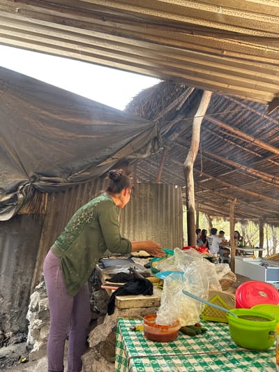The image depicts an outdoor food preparation and dining area in San Miguel Atlapulco, Mexico. In the foreground, a woman with dark hair tied in a bun, wearing an olive green long-sleeved shirt with an intricate pattern on the back and light purple leggings, stands facing right. Her hands are positioned over a green bowl on a stone countertop, appearing to be shaping dough. A dark griddle and a black cloth are visible on the counter. To her right, a lower table is covered with a green and white checkered tablecloth, holding a terracotta-colored bowl with red salsa, a light blue spoon, a green plastic bowl with green salsa, and several small green vegetables. Multiple clear plastic bags filled with various food items are piled on the counter. The background features a rustic structure with a multi-layered roof system composed of horizontal wooden slats, a dark grey tarp, and a thatched section supported by rough wooden poles. Corrugated metal sheeting forms part of the wall behind the woman. To the far right, several individuals are seated at a white rectangular table, engaged in dining and socializing, with beverage bottles visible on their table. A white rectangular cooler or storage unit is present on the far right.