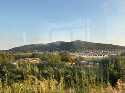 The image depicts a landscape with a village nestled on a hillside, as seen from a moving vehicle. The foreground is dominated by lush green vegetation, blurred due to motion. In the mid-ground, a collection of buildings and industrial structures is visible, including a long, low-rise building with many windows and several white vans. A large, green, forested hill rises behind the structures. The sky above is a clear, pale blue, with some faint reflections from the vehicle's windows visible on the surface. The scene appears to be from the outskirts of La Crau, France.