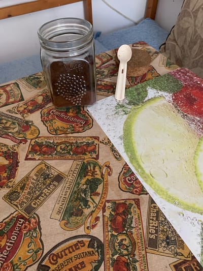 This is a top-down, close-up shot of a table covered with a fabric featuring vintage-style food labels. The fabric has a beige background and is decorated with colorful illustrations and text related to preserved foods, such as tomatoes, chickens, fruits, and vegetables, with some labels in French and Spanish. Prominently featured on the fabric are the words "CharRie's," "Confectionery," "Trois Amis," and "Ballotine de Dinde."

On the fabric, there is a clear glass jar filled with a dark brown substance, likely coffee grounds or spices. Next to the jar is a small, white, plastic spoon, and resting on top of the spoon is a single, round, brown biscuit or cookie. In the upper left corner of the image, a portion of a blue pillow is visible. The lighting suggests it is daytime, but there are no specific weather or strong location cues beyond the fabric's design. The overall impression is one of domesticity and food preparation or presentation.
