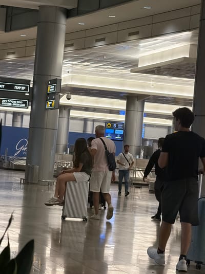 In an airport terminal in Israel, a diverse group of individuals navigate the space. A woman is seated on a silver suitcase near a baggage carousel. To her right, a man wearing a pink shirt and white shorts walks away from the camera, his arm extended as if gesturing or pointing. Further in the background, other travelers are visible, including a man in a white t-shirt and jeans walking towards the camera, and a man in a black t-shirt walking away. The environment is characterized by polished floors, large pillars, and overhead lighting, with signs indicating directions and customs information. The overall scene captures a moment of transit within a modern airport setting.