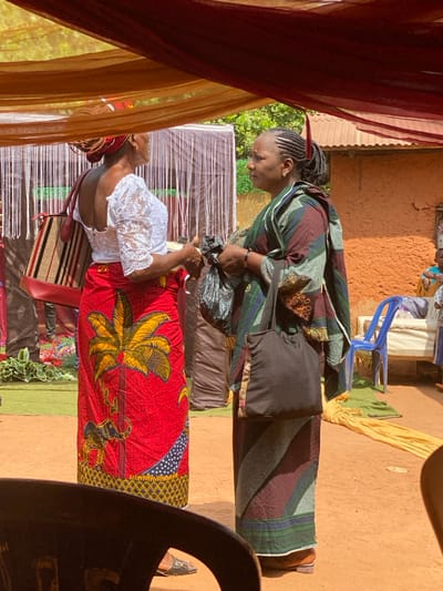The image depicts an outdoor gathering in Ochadam, Nigeria. Two adult women stand facing each other, engaged in conversation on a reddish-brown earth ground. Overhead, a temporary canopy of draped brown and maroon fabric is visible.
The woman on the left, facing right in profile, has dark skin. She wears a white lace short-sleeved blouse and a red patterned wrapper featuring yellow palm trees, blue dots, and other motifs. Her braided hair is covered by a reddish-orange headwrap. She carries a striped woven tote bag with red leather accents on her left shoulder and has her left hand slightly extended towards the second woman. She wears dark flat sandals.
The woman on the right, facing left in profile, also has dark skin. She wears a multi-colored patterned garment, predominantly green, blue, maroon, and brown. Her braided hair is pulled back and adorned with a red feather-like decoration. She holds a black plastic bag in her left hand and carries a dark fabric tote bag over her right shoulder. She wears dark flat shoes.
In the background, a white fringed curtain or decoration is visible, behind which green material covers the ground. To the far right, a building with an orange-brown, textured wall and a corrugated metal roof is partially visible. In front of this building, a blue plastic chair and yellow fabric on the ground are present. Partial figures of other individuals are discernible further back in the scene. A dark chair back is visible in the lower right foreground.