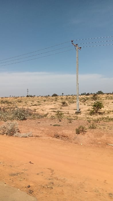 The image depicts an expansive outdoor scene in Barakin Kuru III, Nigeria, characterized by an arid or semi-arid landscape under a clear blue sky with a faint haze near the horizon. In the foreground, a reddish-brown dirt road or track occupies the lower portion of the frame, showing minor surface irregularities and a dark, possibly paved edge at the bottom left. The midground features sparsely vegetated reddish-brown soil, dotted with small, scrubby bushes and low-lying plants displaying green, grey-green, and reddish hues. A single, light grey utility pole, constructed from concrete or a similar material, stands upright slightly right of center, supporting a cross-arm structure from which three power lines extend horizontally across the sky, appearing as dotted lines. A metal utility box is affixed to the lower section of this pole. Further in the distance, towards the left, a taller metallic structure, resembling a communication or transmission tower, is visible against the horizon. The background consists of more distant, indistinct vegetation and trees. No individuals, animals, or explicit actions are present within the frame.