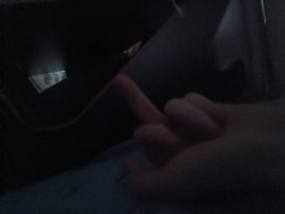 A dark, close-up, slightly blurred image shows a hand with the index finger extended upwards, appearing to touch a dark, curved surface. The background is mostly obscured by shadow, but a hint of light reflects off a shiny, possibly metallic object. The overall scene suggests an indoor environment, likely a personal space, with a low light setting. There is no visible indication of the location in Shipunovo, Russia.
