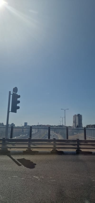 The image shows a traffic light on the left, casting a shadow on the asphalt road. In the background, a highway is visible with cars driving on it. There are streetlights along the highway, and buildings, including a tall, modern structure, are in the distance. The sky is clear and blue with the sun shining brightly, creating lens flare in the upper left corner. The scene appears to be captured from a bridge overlooking the highway. This is a view of the highway crossing into or out of Hod HaSharon, Israel, likely on Road 531 or a similar major artery.