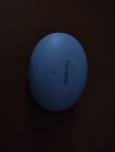 The image shows a light blue, oval-shaped Huawei earbud case resting on a dark brown surface. The Huawei logo is visible vertically on the right side of the case. The case has a speckled texture. The scene appears to be indoors. There is no indication of the location being in Дудзіцкі сельскі Савет, Belarus.