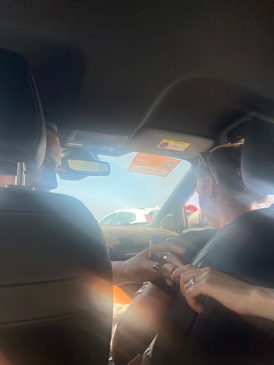 The image captures the interior of a vehicle, looking forward from a rear seat, under bright daylight in Parikia, Greece. Two individuals occupy the front seats. The driver, a male, is seated on the left, visible from behind a dark headrest with red stitching. His left hand, adorned with a ring, is extended towards the center. The front passenger, a female, is seated on the right, also visible from behind a similar headrest. She wears sunglasses on her head and earrings, and multiple rings are visible on her left hand. Her right hand holds a small, cylindrical object of light-colored, possibly wooden material, featuring metallic accents. The male's left hand is engaged with this object, their fingers interlocked as if in the act of opening or closing it. Through the front windshield, a clear blue sky is visible above the rear of a white vehicle and a dark-colored vehicle, indicating an outdoor setting, likely a road or parking area. The vehicle's sun visor on the passenger side displays an "AIRBAG" warning label and a rectangular orange sticker with Greek text, identifying it as from the "Hellenic Republic / Ministry of Citizen Protection / Police" with "SOS" and the emergency number "100." Sunlight causes noticeable lens flares within the cabin.