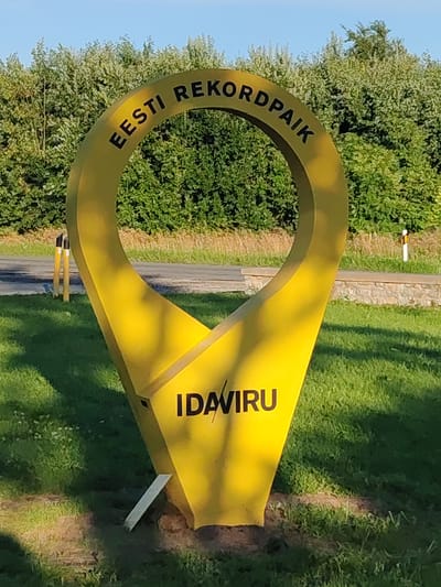 The image shows a bright yellow, pin-shaped monument in Valaste, Estonia. The monument is large and appears to be made of metal. The text "EESTI REKORDPAIK" is written in black letters in a circle around the top opening of the pin. Below the opening, the word "IDAVIRU" is also written in black letters. The monument is set on a grassy area, with a road and trees visible in the background. There are some yellow and white posts along the side of the road. The sky is clear and blue. The lighting suggests it is daytime.