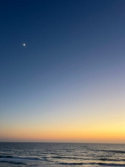 A crescent moon shines in a dark blue sky that transitions to a gradient of pale blue, yellow, and orange. A strip of textured gray, possibly water, occupies the right edge. The scene suggests a time near sunrise or sunset.