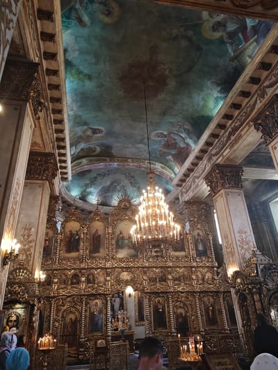 The image depicts the interior of the Raifa Monastery in Mestechko Raifa, Russia, focusing on its opulent iconostasis. A large, ornate golden iconostasis dominates the central portion of the frame, featuring multiple tiers of icons set within gilded frames. The icons themselves are religious paintings, many depicting figures with halos. A large, multi-tiered crystal chandelier hangs from the ceiling above the iconostasis, illuminated with numerous candles. The ceiling is adorned with vibrant frescoes depicting religious scenes and figures, with a predominantly green and blue color palette. Elaborate golden architectural details, including columns and decorative moldings, frame the ceiling frescoes and the sides of the iconostasis. Several lit candles are visible on stands in front of the iconostasis, and a few individuals, mostly seen from behind or the side, are present in the foreground and midground, suggesting a religious service or visitation. Their attire appears modest, with some women wearing headscarves. The overall impression is one of grandeur, religious devotion, and rich artistic heritage.