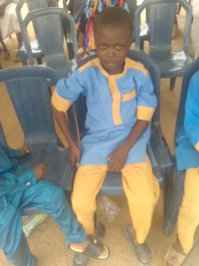 The scene depicts a young male individual, approximately 6-8 years old, seated in a blue plastic chair. The location is Ijoko, Nigeria. He wears a light blue long-sleeved tunic with yellow trim on the collar, sleeves, and a vertical strip down the front, paired with matching yellow trousers. His feet are clad in dark-colored slip-on sandals. He maintains a direct gaze towards the camera.
To his immediate left, another individual, likely a child, is partially visible, also seated in a similar blue plastic chair and wearing a patterned teal-blue garment with gold buttons, along with dark slip-on sandals. Behind the central figure, multiple empty blue plastic chairs are arranged. Blurred figures of other individuals are visible in the background, also seated or standing, some wearing traditional attire. The ground surface appears to be packed dirt or sand. No explicit interactions are observed between the individuals in the immediate foreground; they are positioned in proximity.