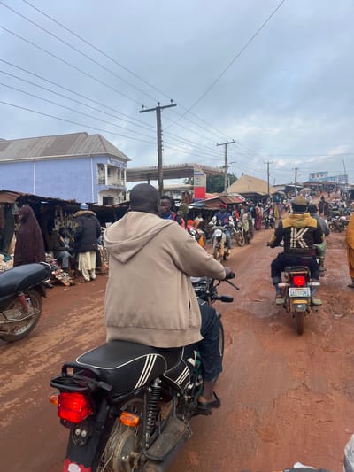 The image depicts a busy street scene in Gembu, Nigeria, likely during the day given the bright, overcast sky. The primary activity is a congregation of motorcycles and people, suggesting heavy traffic or a market day.
The foreground features the back of a person on a motorcycle, with their legs and feet visible. The motorcycle itself is dark with white stripes on the seat and a prominent red taillight. The ground is a reddish-brown, indicative of dirt or unpaved roads common in the region.
In the mid-ground and background, numerous other motorcycles are parked or in motion, with many people, some wearing traditional clothing, gathered around. There are stalls and shops lining the street, suggesting a commercial area. One building has a blue facade with white architectural details. Overhead, utility poles and wires are visible, characteristic of a developed area.
There is some visible text on a license plate of a motorcycle in the mid-ground, reading "GAS 5224 U" and a star emblem. Other signs are too blurry to read clearly, but their presence indicates ongoing commerce. The overall atmosphere appears lively and active, with a sense of community engagement in a marketplace setting. The overcast sky suggests potentially humid or rainy weather.