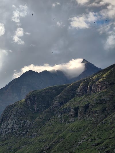 The image captures a dramatic mountain landscape under a cloudy sky, with several birds in flight. In the foreground, a steep, green, and rocky mountainside dominates the lower portion of the frame. The terrain is rugged, with visible rock formations interspersed with patches of vegetation.

Further back, a darker, more imposing mountain peak rises, partially obscured by a bright, white cloud bank that wraps around its summit. The sky above is a mix of dark grey storm clouds and patches of brighter, illuminated sky, suggesting dynamic weather conditions.

Several small, dark silhouettes of birds are visible in the sky, scattered across the frame. They appear to be in mid-flight, adding a sense of movement and scale to the vastness of the mountains and sky. The lighting suggests it may be late afternoon or early evening, with shafts of light breaking through the clouds to illuminate certain parts of the mountains. The scene evokes a sense of wild, natural beauty. This location is identified as Yari-Khoksar, India.