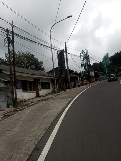 The image depicts a street scene in Bangtsho, Bhutan, under an overcast sky. The viewpoint is from a low angle, looking forward along an asphalt road that curves slightly to the right and descends. A solid white line delineates the left edge of the road, bordering a cracked concrete sidewalk.
On the left, a row of low-rise buildings lines the street. The nearest structure has white-painted lower walls, a dark weathered roof, multiple windows, a door, and a circular blue sign affixed to its facade. Further down the street, subsequent buildings exhibit varied materials, including corrugated metal, and some display commercial signage with text such as "MIE AYAM," "BASO RAHAYU," and "WARUNG RASA KITA." Multiple wooden utility poles, heavily strung with black electrical wires, and a metallic streetlight pole are prominent on this side. Further along, several large, empty green or dark metal billboard frames rise above the rooftops.
On the right side of the road in the mid-ground, a dark-colored SUV is visible. The background on the right is dominated by a densely forested hillside. No people are visible in the scene. The sky is uniformly grey and cloudy.