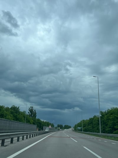 The image shows a highway with a gray, overcast sky. Trees line both sides of the road, and there are sound barriers on the left. Several cars are visible on the highway, and there are streetlights along the road. The overall atmosphere is moody and perhaps suggestive of impending rain. The location is identified as Sokolivka, Ukraine.