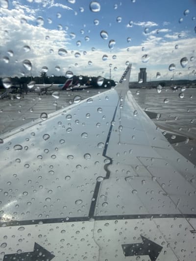The image shows a close-up view of an airplane wing with water droplets on the window, taken from inside the aircraft. The wing is white with a dark stripe along its edge. The word "RYANAIR" is visible on the tail fin. In the background, there is an airport tarmac with other airplanes and airport buildings. The sky is cloudy with patches of blue. The scene appears to be at John Paul II International Airport Kraków–Balice, located in Balice, Poland. No people are visible in the image. The overall impression is a typical airport scene during or after rain.