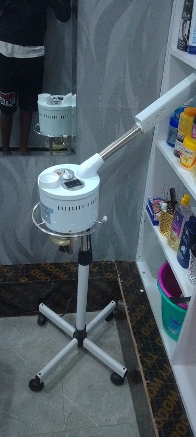 The image depicts an indoor scene, likely within a salon or beauty treatment establishment in Matathani ward, Kenya.

In the foreground, a white, mobile facial steamer is positioned centrally. It consists of a cylindrical main unit with ventilation slots, a control panel on its top surface featuring a rotary dial and two rocker switches, and an adjustable, articulated arm ending in a nozzle. This unit is mounted on a height-adjustable stand with a five-star base equipped with black caster wheels. A circular chrome-plated rail surrounds the upper portion of the steamer unit.

To the left, a vertical rectangular mirror is affixed to a grey wall patterned with subtle wavy designs. The mirror reflects the lower torso and legs of an individual wearing a dark top (partially visible), a white undershirt, and black shorts with a white graphic on the left leg. The individual's skin appears dark. The mirror also reflects the upper section of the steamer.

To the right of the steamer, a white, open shelving unit is visible. The top shelf holds multiple plastic bottles of various colors and sizes, appearing to be hair or beauty products, with "Bamsi Hair Food" partially legible on a yellow bottle. The middle shelf contains additional bottles, including a clear one with purple liquid and a dark blue one, along with several dark brushes and a small golden metallic container. The bottom shelf holds a two-tone green and pink plastic bucket, partially filled with dark items.

The floor beneath the steamer and extending to the left is covered with light grey square tiles exhibiting a subtle dark veining pattern. To the right, beneath the shelving unit, the floor transitions to a dark plywood or particle board material displaying repeated white text, primarily "WOOD" and "TY" in a grid formation.