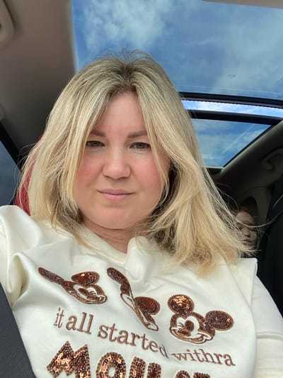 In a car with a sunroof, a blonde woman with fair skin and light green eyes is looking directly at the camera. She has shoulder-length, wavy blonde hair. She is wearing a cream-colored sweatshirt with a graphic print. The print features three Mickey Mouse heads outlined in sequins and the text "it all started withra" and "Mom" also outlined in sequins. In the background, a child with reddish hair is visible in the back seat. The sunroof shows a blue sky with scattered clouds. The scene appears to be outdoors, likely in a suburban or rural area, given the clear sky and natural light. There is no specific indication of the city of Поварёнки, Russia.
