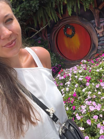 A woman with brown hair and green eyes smiles at the camera in a selfie taken outdoors. She is wearing a white off-the-shoulder top and a black crossbody bag. Behind her, a circular red door with a wreath is visible, set into a whimsical structure that resembles a hobbit hole. Lush green foliage and colorful pink and white flowers surround the scene. The location appears to be a themed garden or attraction, potentially in Da Lat, Vietnam, given the style of the garden and the resemblance to attractions found in the city known for its flower gardens and European-inspired architecture.