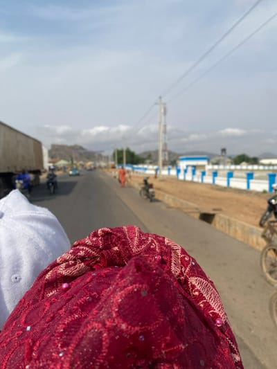 The image depicts an outdoor, daylight scene along a paved roadway in Abobo, Nigeria, observed from a low vantage point, likely from within a vehicle or atop a motorcycle. In the immediate foreground, out of focus, are the upper portion of a white fabric object, resembling a cap or head covering, and a prominent dark red, intricately patterned fabric, possibly lace or embroidered, adorned with small, reflective elements like sequins or beads, dominating the lower right quadrant.
The road stretches into the distance, populated with various vehicles and individuals. On the left side of the road, a large, light-colored cargo truck is visible, followed by at least two individuals operating motorcycles and several passenger vehicles, one of which appears light blue. On the right side of the roadway, a raised dirt shoulder borders a prominent white wall accented with blue pillars or trim, possibly belonging to a compound or facility. A person wearing an orange garment is walking on the right side of the road, and another individual is seen on a bicycle further along, near the white and blue wall. Utility poles with overhead wires line the road, and a tall cellular communication tower is visible in the mid-distance on the right. The background features a range of hills or low mountains under a partly cloudy sky.