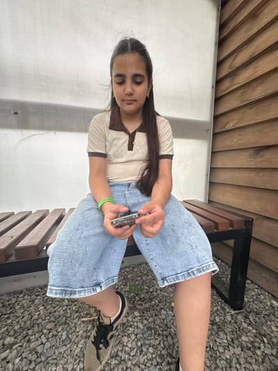 A young girl is sitting on a wooden bench in Zaliznychne, Ukraine. The bench is outdoors, with a pebbled ground and a wooden wall behind it. The girl is wearing a light brown polo shirt with a dark brown collar and trim, and light blue wide-leg jeans. She is holding a small silver object, possibly a phone or music player, in her hands. She has long dark hair and is looking down at the object. A green bracelet is visible on her left wrist. The background consists of a white translucent wall on the left and a horizontally slatted wooden wall on the right.