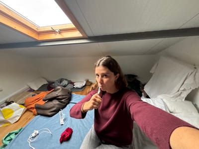 The scene depicts an interior room, specifically an attic or loft space, located in Montemor-o-Novo, Portugal. A female subject, seen from the chest up, occupies the center-right of the frame, engaged in brushing her teeth with a white toothbrush while simultaneously taking a selfie with her extended right arm. She has brown hair, pulled back, and is wearing a dark red long-sleeved sweater and light-colored pants. A silver earring is visible in her left ear. The room features a sloped white ceiling, with a wooden-framed rectangular skylight at the top center revealing an overcast sky. A dark horizontal beam traverses the upper half of the room. The floor is partially covered by a blue mattress or pad, upon which a folded brown garment, an orange textile, a white charging cable with an adapter, a red fluffy hair accessory, and a clear plastic item are present. To the left, a white pillow, a white and yellow patterned shopping bag, and various other textiles in green and grey hues are visible on the floor against the wall. On the right, a bed with white sheets and a white patterned cover is partially obscured by the subject.