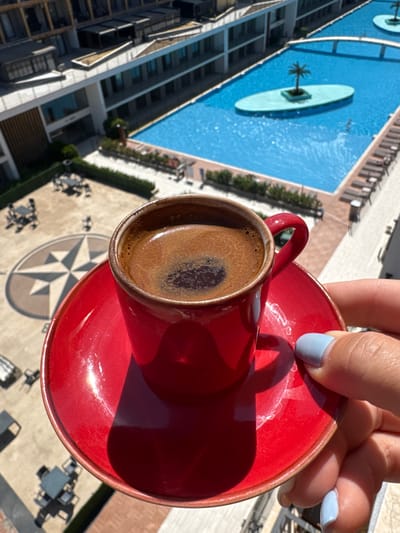 An aerial, eye-level shot captures a red espresso cup filled with dark coffee, set on a matching saucer. The cup is held by a hand with light blue nail polish. In the background, a large blue swimming pool with an artificial island featuring a palm tree is visible, adjacent to a multi-story building. Below, an outdoor seating area with tables and chairs is laid out on a paved surface with a compass rose design. This scene is located at the Ornekkoy Aqua Beach Club in Çiğli, Turkey.