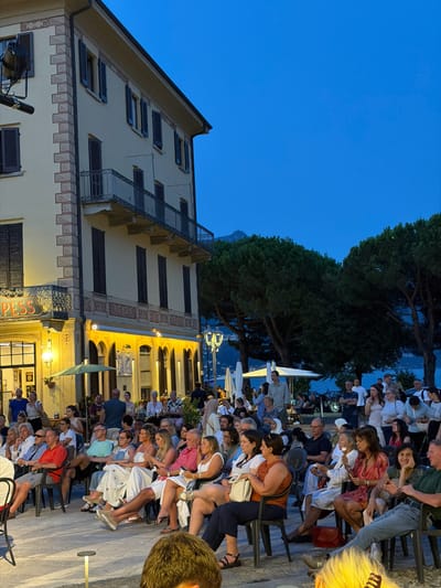A crowd of people are seated outdoors in chairs, facing away from the camera, watching an event. The scene appears to be in Loveno, Italy, given the architecture and the presence of a body of water in the background. The building behind the crowd features a light-colored facade with arched windows and balconies, illuminated by warm lights. Green trees are visible on the right side of the image, and a body of water, likely a lake, is in the distance under a twilight sky. The people are dressed in casual summer attire, and many are looking towards the unseen event.