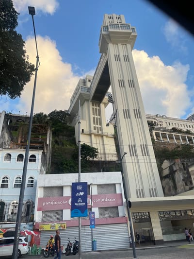 The image presents a street-level view of the Elevador Lacerda in Salvador, Bahia, Brazil, specifically from the Cidade Baixa (Lower City). The central subject is the multi-story, cream-colored Art Deco elevator complex, comprising two vertical towers connected by an overhead horizontal bridge, facilitating transit to the Cidade Alta (Upper City). The visible tower features repetitive vertical fluted designs and recessed rectangular window groupings.
To the left of the elevator, a light blue and white building displays "panificação e confeitaria" (bakery and confectionery) lettering on its facade, with its street-level entrance secured by corrugated metal shutters. Several blue and white banners, advertising "SALVADOR PREFEITURA" and other civic initiatives, are mounted on tall, black streetlights positioned along the foreground.
On the paved street, a white car is parked on the far left, partially obscured. Two individuals are visible walking on the sidewalk in the midground, near three parked motorcycles. The background shows a steep, vegetated cliff face rising between the lower and upper city levels, with additional building structures, including an ornate white building, visible towards the upper right. The sky is predominantly blue with scattered white clouds, some exhibiting a yellowish tint from the ambient lighting conditions.