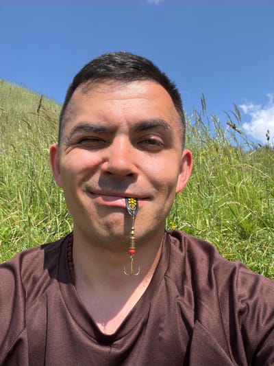 A man is captured in a close-up, selfie-style photograph, with his face filling most of the frame. He has short, dark hair and is looking towards the camera with his left eye squinted. A fishing lure, featuring a gold-colored treble hook, is held between his lips. The lure has a black and yellow patterned front, with metallic beads and a red accent. The man is wearing a brown, long-sleeved shirt and a beaded necklace. The background consists of tall green grass and a bright blue sky with a few scattered clouds, suggesting an outdoor, possibly rural or natural setting. The location of Podshivalovo, Russia, is not discernible from the visual information present in the image.