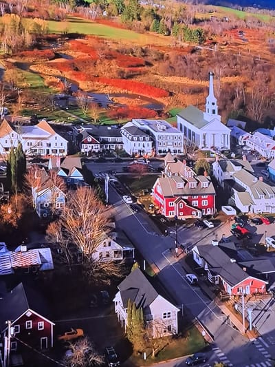 This is an aerial, high-angle photograph of a residential neighborhood during autumn. The image is oriented vertically, with the top of the frame showing a landscape of trees displaying vibrant fall foliage in hues of red, orange, and yellow, interspersed with green grass. The lower portion of the frame is dominated by houses and streets.
The houses are varied in architectural style and color, with many featuring peaked roofs in shades of gray, brown, and white. Several houses are painted red with white trim. Streets are visible, with cars parked along them and some driving. There are also signs of infrastructure such as streetlights and sidewalks.
Notable details include the lush, colorful autumn foliage, suggesting a time of year in late autumn. The lighting appears to be daytime, likely afternoon given the shadows cast by the houses and trees. There are no people clearly visible in the image, and the activity appears to be static, depicting a residential scene. No visible text is present in the image. The context provided of "Cēsis, Latvia" places the scene in a European setting during the fall season.