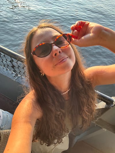 A woman with long, brown, wavy hair and tortoise-shell sunglasses is taking a selfie on a boat. She is wearing a light olive-colored sleeveless top and a white beaded necklace. Her right hand is holding the arm of her sunglasses, and the sun is hitting her face and arm. The background shows rippling water and a dark railing with a mesh pattern. The image appears to have been taken in the city of Verhnij Uslon, Russia.