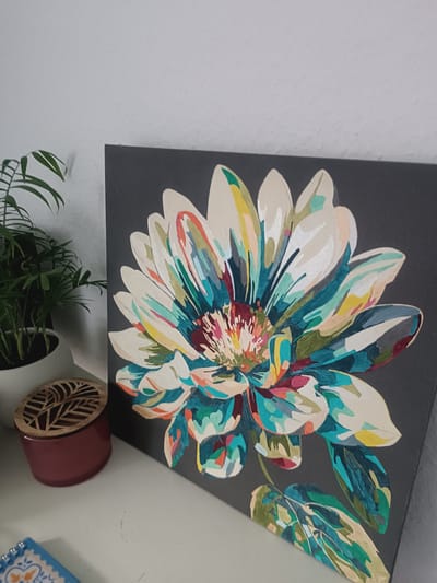 The image displays a close-up, angled shot of a painting of a vibrantly colored flower against a dark gray background. The flower has large, broad petals with a central cluster of delicate, multi-hued stamens. The petals are painted with broad strokes of white, cream, teal, yellow, pink, and green, giving the flower a dynamic and textured appearance. The painting is positioned on a white shelf next to a potted green plant. To the left of the painting is a small, reddish-brown cylindrical container with a decorative wooden lid featuring a leaf pattern. A portion of a small, blue and white tiled object is visible in the lower left corner. The overall scene is indoors, with a white wall visible in the upper left background. There are no people present in the image. The location of this scene is not specified within the image itself.